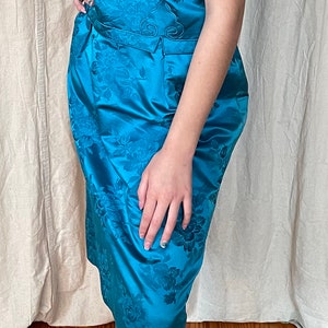 May include: A teal blue dress with a floral pattern and short sleeves. The dress has a fitted silhouette and a cinched waist. The model is wearing clear heeled sandals.