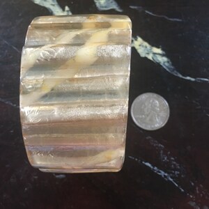 May include: A clear, ribbed bangle bracelet with a swirl of white and brown inside. The bracelet is about the size of a quarter.