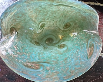 May include: A large, green glass bowl with a swirling, marbled pattern in shades of gold and white. The bowl has a wide, shallow shape and a slightly rippled edge.