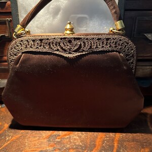 May include: A brown velvet handbag with a gold clasp and a delicate lace trim. The handbag has a top handle and a rounded shape.