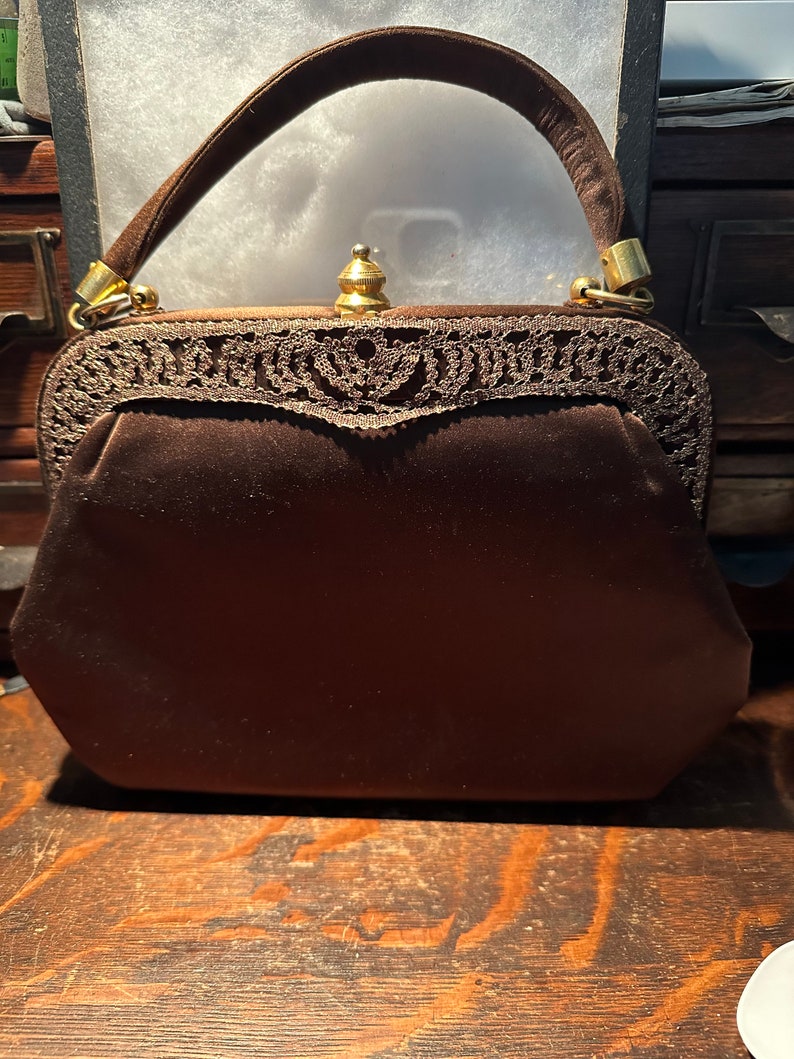 May include: A brown velvet handbag with a gold clasp and a delicate lace trim. The handbag has a top handle and a rounded shape.