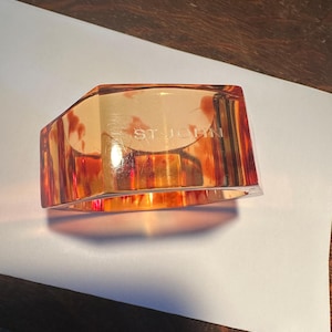 May include: An amber-colored, geometric bracelet with the text "ST. JOHN" visible. The bracelet has a faceted design, reflecting light and creating a warm, glowing effect. The piece is set against a white surface.