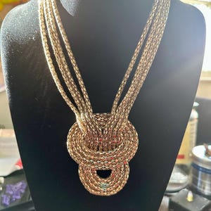May include: A gold-toned necklace with multiple strands cascading into a knotted design. The necklace is displayed on a black mannequin, showcasing its intricate woven pattern and overall structure. The necklace is a statement piece.