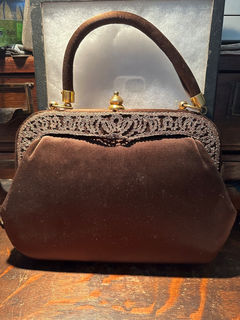 May include: A brown velvet handbag with a gold metal clasp and a decorative lace trim. The handbag has a brown velvet handle.