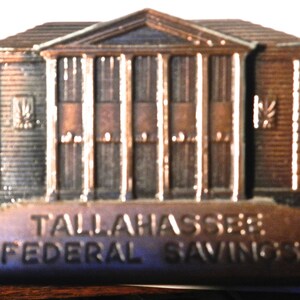 May include: A bronze-colored metal bank shaped like a building with a sign that reads "Tallahassee Federal Savings".