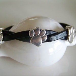 May include: A black leather bracelet with silver paw print charms. The bracelet is braided and has three charms.