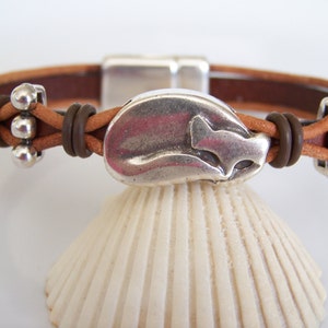 May include: A brown leather bracelet with a silver metal charm shaped like a fish. The bracelet has a magnetic clasp and is decorated with small silver beads.