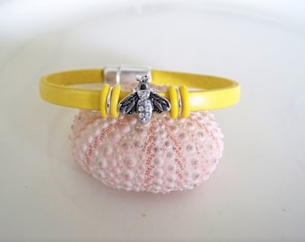 May include: A yellow leather bracelet with a silver bee charm. The bracelet has a magnetic clasp and is accented with silver rings.
