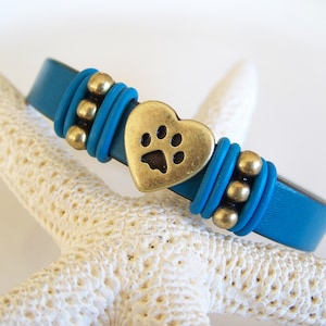 May include: Teal leather bracelet with a gold heart charm featuring a paw print. The bracelet is accented with gold beads.