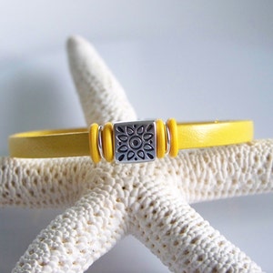 May include: A yellow leather bracelet with a silver charm featuring a sun design. The bracelet is on a white starfish.
