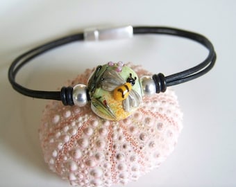 May include: A black leather bracelet with a silver clasp and a glass bead featuring a bee and flowers. The bead is green, yellow, and white.