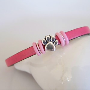 May include: A pink leather bracelet with a silver paw print charm and pink beads.