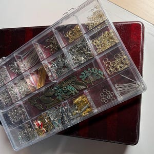 May include: A clear plastic organizer box filled with various metal jewelry making supplies, including gold and silver jump rings, eye pins, earring hooks, and assorted leaf-shaped charms in bronze, copper, and green patina finishes.