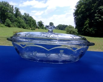 May include: Clear glass serving dish with a lid. The dish has a scalloped edge design with floral patterns and a decorative handle on the lid. The dish is sitting on a blue surface, with a green background.