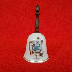 May include: White ceramic bell with a wooden handle. The bell features a painting of Norman Rockwell's "The Artist's Studio" with the text "Norman Rockwell 1961" at the bottom.