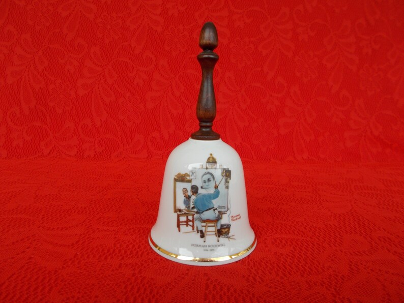 May include: White ceramic bell with a wooden handle. The bell features a painting of Norman Rockwell's "The Artist's Studio" with the text "Norman Rockwell 1961" at the bottom.