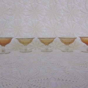 May include: Five vintage amber-colored glass dessert dishes. Each dish has a round bowl with vertical ribbed detailing and a clear stem and base. The dishes are arranged in a row against a white lace background.