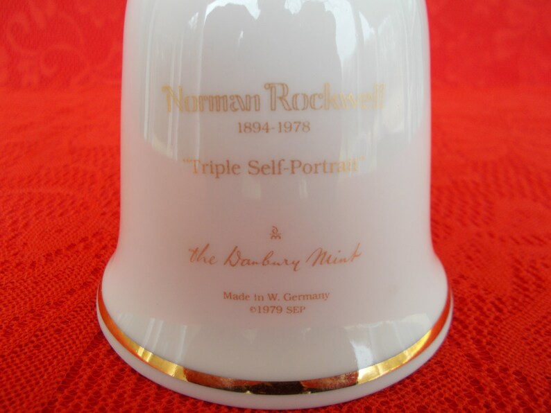 May include: White porcelain bell with a gold trim. The bell features a gold inscription that reads "Norman Rockwell 1894-1978 Triple Self-Portrait" and "The Danbury Mint Made in W. Germany &copy;1979 SEP".