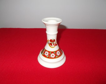 May include: A white ceramic candlestick holder with a flared base and a decorative band featuring red, white, and green floral and holiday-themed designs. The candlestick has a cylindrical body and a top designed to hold a candle.
