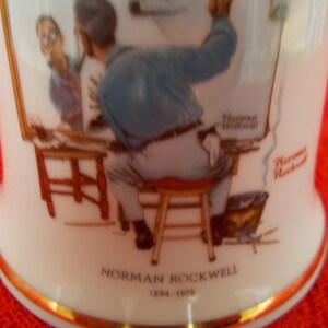 May include: A white ceramic mug with a gold rim features a painting of Norman Rockwell's "Triple Self Portrait". The mug has the artist's name and dates "Norman Rockwell 1894-1978" printed on the bottom.