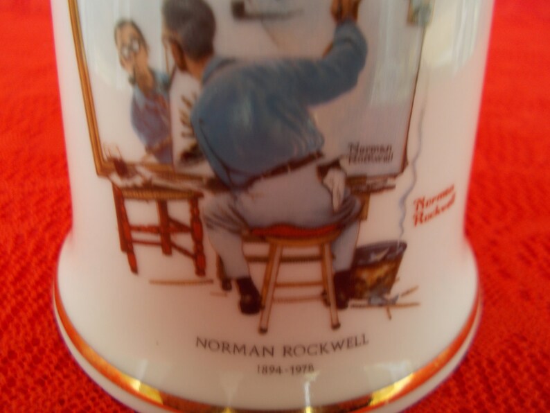 May include: A white ceramic mug with a gold rim features a painting of Norman Rockwell's "Triple Self Portrait". The mug has the artist's name and dates "Norman Rockwell 1894-1978" printed on the bottom.