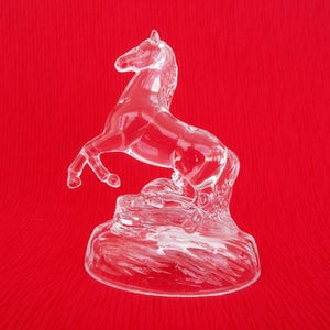 May include: A clear glass horse figurine with a raised tail and mane, standing on a textured base. The figurine is on a red background.