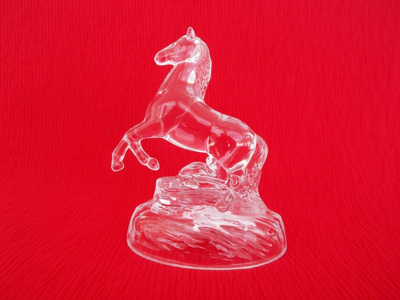 May include: A clear glass horse figurine with a raised tail and mane, standing on a textured base. The figurine is on a red background.