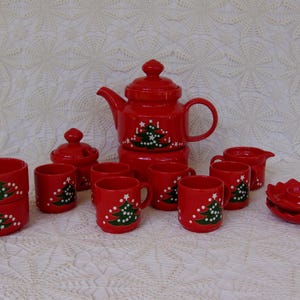 May include: A red ceramic Christmas tea set with a teapot, cups, sugar bowl, creamer, and a serving dish. Each piece is decorated with a green Christmas tree design and white accents. The set is displayed on a white lace tablecloth.