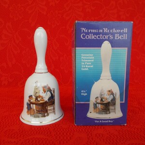 May include: White porcelain bell with a gold trim, featuring a Norman Rockwell illustration of a man and a boy building a ship. The bell is 6 1/2 inches high and comes in a blue box with the text "Norman Rockwell Collector's Bell" and "For A Good Boy".