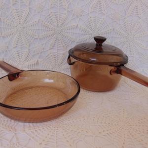 May include: A vintage amber-colored glass cookware set, including a frying pan and a saucepan with a lid. The pan has a long handle, and the saucepan has a handle and two side grips. The set is made of transparent glass.