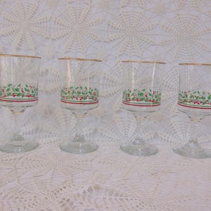 May include: Four clear wine glasses with gold rims and a festive green and red holly berry design around the base. The glasses are displayed on a white lace tablecloth, creating a vintage aesthetic.
