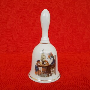 May include: White porcelain bell with a gold trim. The bell features a painting of a man and a boy sitting at a table with a ship model. The text on the bell reads "The Skipper's Mate".