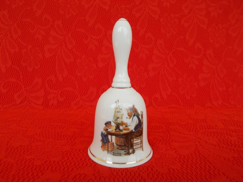 May include: White porcelain bell with a gold trim. The bell features a painting of a man and a boy sitting at a table with a ship model. The text on the bell reads "The Skipper's Mate".
