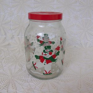 May include: A clear glass jar with a red lid, decorated with colorful snowmen in winter attire. The snowmen are wearing hats, scarves, and ice skates. The jar is likely for storing cookies or other treats.