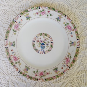 May include: A white ceramic dinner plate with a floral design in shades of pink, blue, and green. The plate has a gold rim and a central design of a vase with flowers.