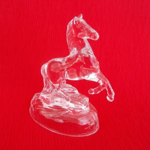 May include: A clear glass horse figurine standing on a base. The horse is rearing up on its hind legs with its front legs raised in the air. The figurine is on a red background.