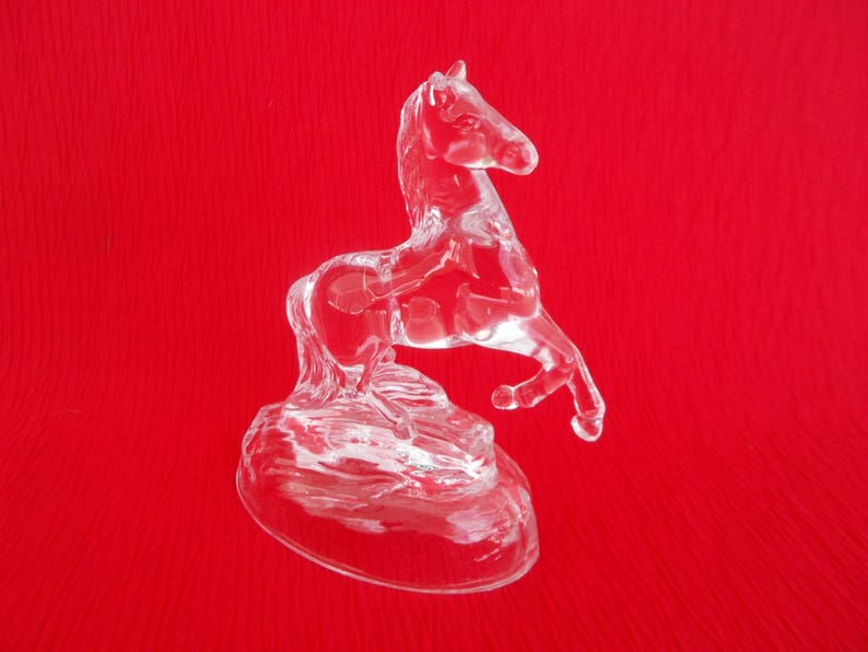 May include: A clear glass horse figurine standing on a base. The horse is rearing up on its hind legs with its front legs raised in the air. The figurine is on a red background.