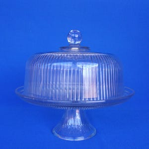 May include: A clear glass cake stand with a domed lid. The stand has a pedestal base and a round plate. The lid has vertical ridges and a small knob on top. The stand is on a blue background.