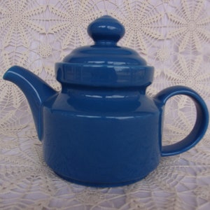 May include: A blue ceramic teapot with a lid and a handle. The teapot is sitting on a white crocheted tablecloth.