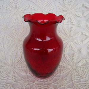 May include: A vibrant red glass vase with a ruffled rim and a dark trim. The vase has a classic shape and a textured surface. This decorative vase is suitable for floral arrangements or as a standalone accent piece.