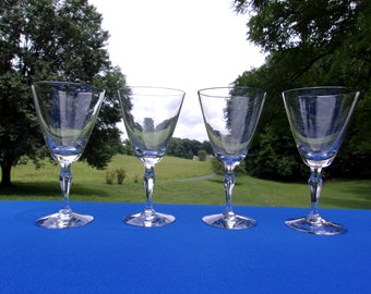 May include: Four clear glass wine glasses with a stemmed base and a round bowl. The glasses are arranged in a row on a blue tablecloth.