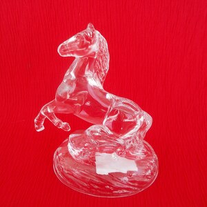 May include: A clear glass horse figurine with a raised tail and mane. The horse is standing on a circular base.