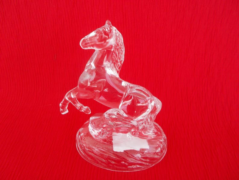 May include: A clear glass horse figurine with a raised tail and mane. The horse is standing on a circular base.