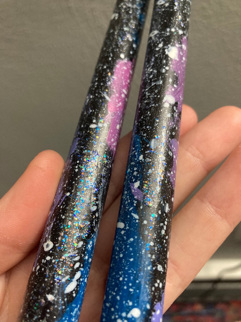 Puede incluir: Un par de baquetas negras con un dise&ntilde;o de galaxia. Las baquetas est&aacute;n pintadas con pintura salpicada azul, p&uacute;rpura y blanca y tienen detalles de purpurina plateada.