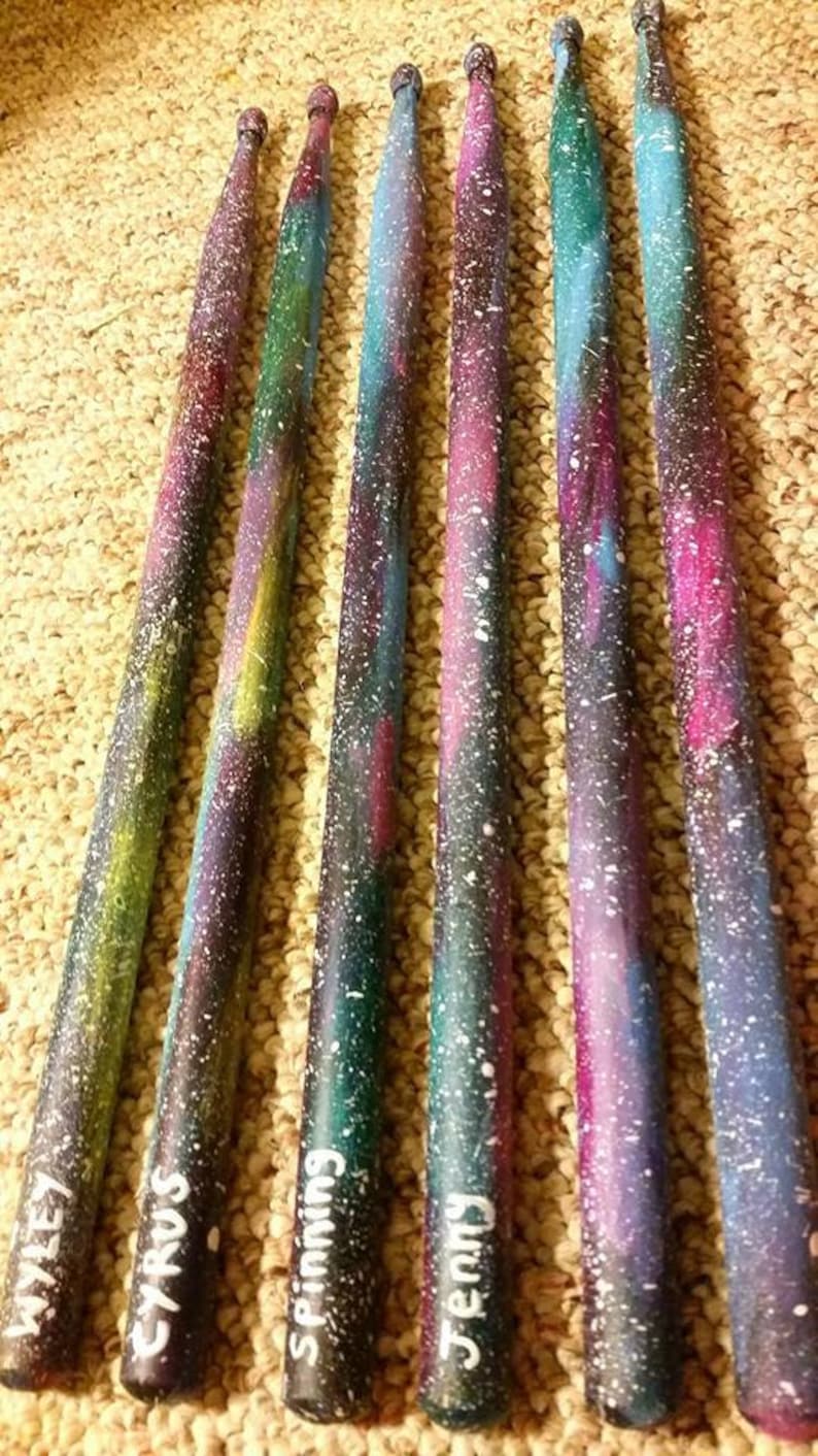 Puede incluir: Seis baquetas de madera pintadas con un dise&ntilde;o de galaxia. Las baquetas est&aacute;n pintadas en tonos de azul, morado y rosa con estrellas blancas. Cada baqueta tiene un nombre escrito en ella: Miley, Cyrus, Spinning, Jenny y otros dos nombres que no son visibles.