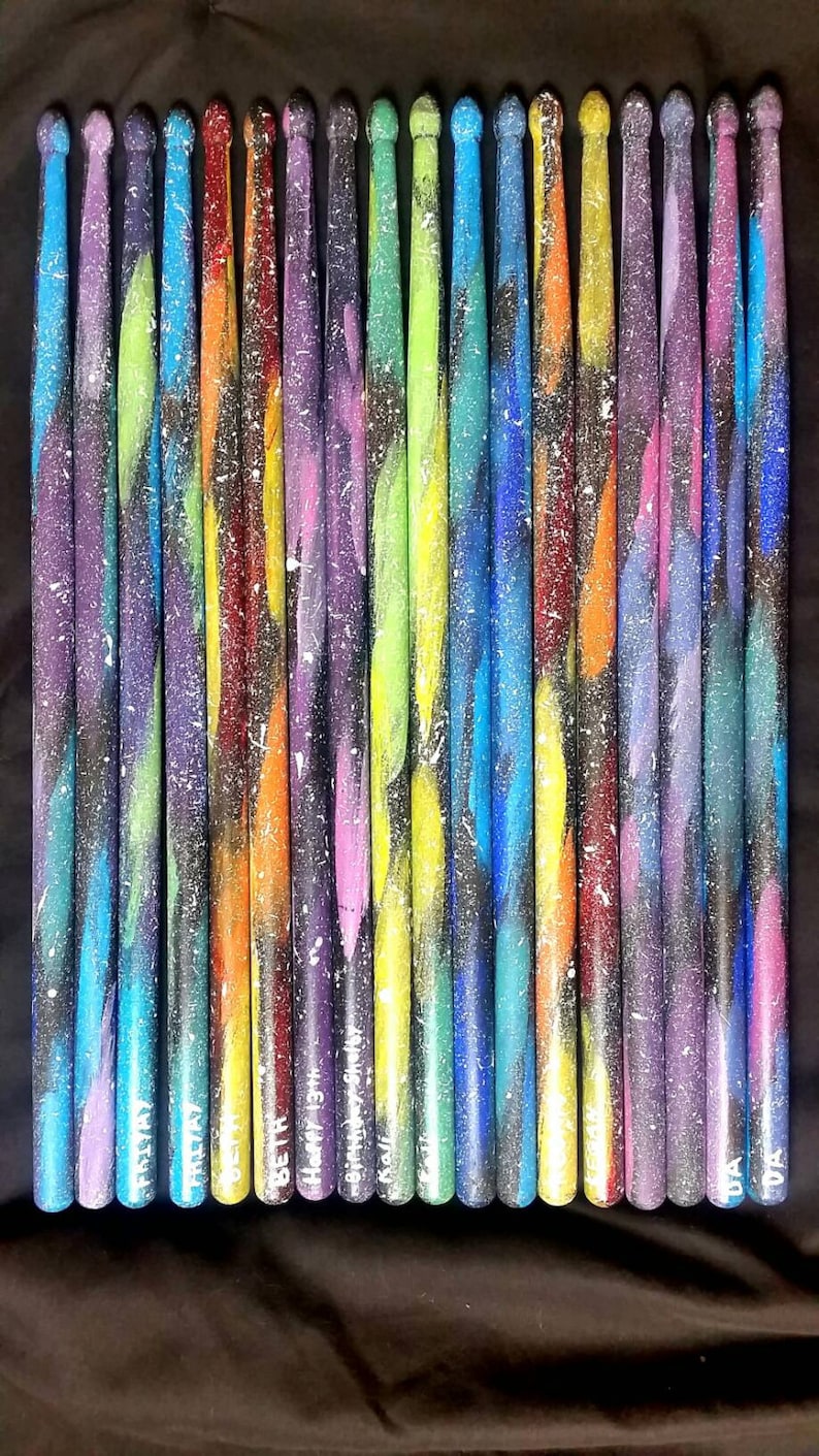 Puede incluir: Veintidos baquetas de madera coloridas con un dise&ntilde;o de pintura inspirado en la galaxia. Cada baqueta est&aacute; personalizada con un nombre o un mensaje, incluyendo "Happy 13th Birthday Sister", "Beth", "Trisha" y "Lisa".