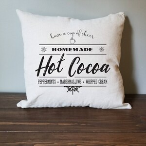 May include: White throw pillow with black text that reads "have a cup of cheer, * HOMEMADE * Hot Cocoa PEPPERMINTS MARSHMALLOWS WHIPPED CREAM"
