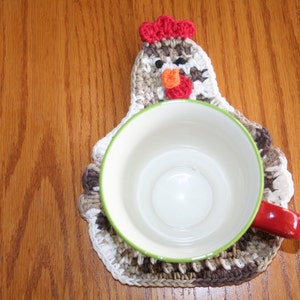 以下が含まれることがあります： 白、茶色、赤のデザインの、鶏の形をしたかぎ針編みのコースター。緑色の縁取りのある白いマグカップがコースターの上に置かれています。