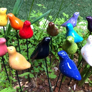Puede incluir: Una colección de 13 figuritas de pájaros de cerámica de colores con pintura moteada, cada una posada en una estaca de metal negro. Los pájaros están dispuestos en un entorno de jardín.
