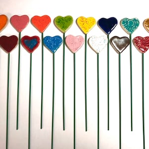 Pode incluir: Um conjunto de 15 estacas de jardim de cerâmica em forma de coração em várias cores e padrões. As estacas são feitas de metal verde e têm uma parte superior de cerâmica em forma de coração. Os corações são decorados com diferentes cores, padrões e texturas.